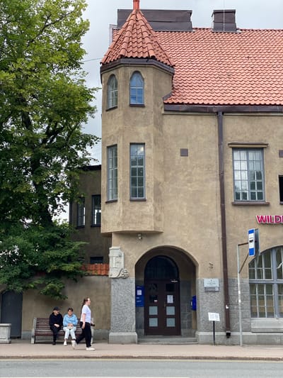 The image shows a street view in Sortavala, Russia, focusing on the exterior of a tan-colored building with a red tile roof and a prominent turret. In front of the building, there is a sidewalk with a bench where two individuals are seated, and another person is walking past them. The building features arched doorways, multiple windows, and a sign that appears to read "WILD." A bus stop sign is visible on the right side of the image. Lush green trees are present on the left, partially obscuring the building. The sky is overcast.