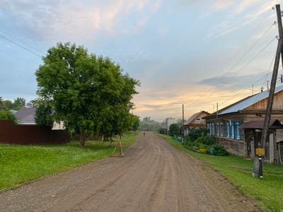 The image depicts a rural road in the city of Baklushi, Russia, under a cloudy sky transitioning from dawn to day. The road is unpaved and appears to be made of dirt and gravel, with tire tracks visible. On the left side of the road, there is a lush green grassy verge bordered by a brown corrugated metal fence. A large, leafy green tree stands prominently on the left side. Beyond the fence, parts of a house with a dark roof are visible.
On the right side of the road, several wooden houses are situated. These houses have wooden facades and some feature blue-trimmed windows. A wooden utility pole with insulators and wires stands near one of the houses. The grass on the right verge is also green, with some flowering plants visible in front of one of the houses.
Further down the road, a single person is walking away from the camera. In the distance, more houses and trees are visible, with a hazy, misty atmosphere suggesting it is early morning. The overall scene conveys a sense of a quiet, rural settlement.