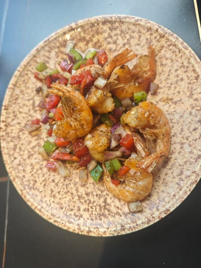 A close-up, overhead shot captures a speckled ceramic plate filled with seasoned shrimp and diced vegetables. The shrimp, with their tails still attached, are a reddish-orange hue and appear to be pan-seared, glistening with oil and spices. Interspersed among the shrimp are cubes of red bell pepper, green bell pepper, and red onion, some softened by cooking. The plate itself has a mottled brown and tan pattern. The background consists of a dark, flat surface, possibly a table, with a thin vertical line of light visible on the left side. The scene is indoors, likely a restaurant or home setting. The location in El Puerto de Santa María, Spain, is not discernible from the image itself.