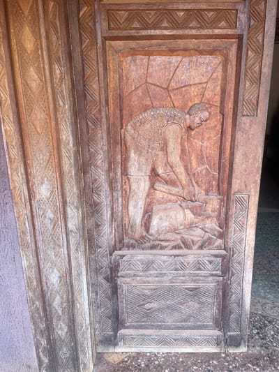 The image showcases a detailed wood carving. A muscular figure, possibly a man, is prominently displayed, lying on his side and appearing to manipulate objects. The figure wears a patterned vest and is set against a background of radiating lines. The carving incorporates geometric patterns along the edges and within the frame. The wood appears aged, with some discoloration and signs of wear.