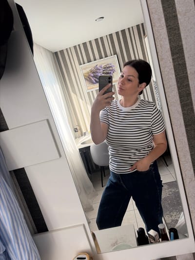 A woman with dark hair tied back takes a mirror selfie in a room. She is wearing a black and white striped t-shirt and dark denim jeans. Her left hand is in her jeans pocket, and her right hand holds a smartphone, capturing her reflection. The background shows a wall with vertical stripes, a framed picture of lavender on the wall, sheer curtains, and a glimpse of furniture. The overall impression is of a domestic setting. The location is identified as Sochi, Russia.