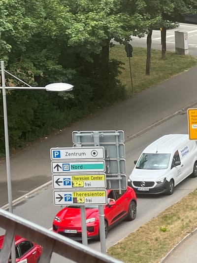 The image shows a street scene in Straubing, Germany, with a focus on traffic signs and vehicles. In the foreground, a set of directional signs are mounted on a metal pole. These signs indicate "Zentrum" (Center), "Nord" (North), "Theresien Center", and "Theresientor" (Theresien Gate), with parking symbols and arrows pointing in various directions. One sign indicates "P 329 frei" (P 329 free) and another "P 53 frei" (P 53 free), suggesting parking availability.
A bright red sports car is parked on the right side of the road, partially obscuring the signs. Its license plate is partially visible as "SR-HS 448E". Further up the road, a white delivery van with "NaturSTEIN Götzfried" written on its side is also visible. Its license plate reads "SRG 1030".
In the background, lush green trees line the left side of the road, suggesting a pleasant outdoor setting. The lighting indicates it is daytime, possibly late morning or early afternoon. The overall impression is of a typical urban street scene in Germany. There are no people clearly visible in the image. The focus is on the road infrastructure and the vehicles present. The image is taken from an elevated perspective, looking down onto the street.
