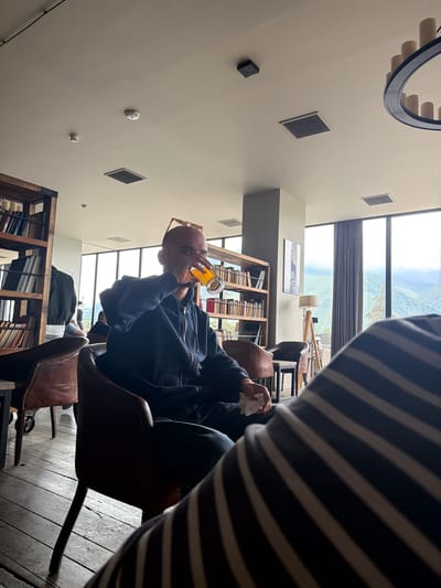 A bald man sits in a leather armchair in a cafe or library in Stepantsminda, Georgia, drinking from a glass of amber liquid. He wears a navy blue hooded jacket and has sunglasses perched on his head. The cafe features wooden floors, a large bookshelf filled with books, and large windows offering a view of a mountainous landscape. To the right, a portion of another person's body, covered in a striped fabric, is visible in the foreground. Other patrons and furniture are present in the background.