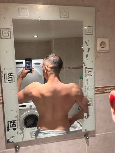A shirtless male is standing in a bathroom in an apartment, taking a picture of himself in a mirror with a smartphone. The mirror is decorative and appears to have some damage. Reflected in the mirror are the man, a white washing machine, and bathroom wall tiles. A power outlet is visible on the wall beside the mirror. The location is possibly Lelystad, Netherlands.