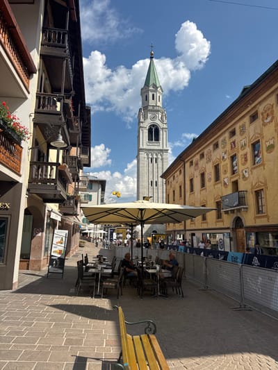 The image depicts a sunlit street scene in Cortina d'Ampezzo, Italy. In the background, a tall, ornate bell tower with a green spire stands prominently against a blue sky dotted with fluffy white clouds. To the right of the tower, a building features a yellow facade adorned with numerous colorful, detailed murals depicting various crests and scenes.
In the foreground, the street is paved with cobblestones, and several outdoor cafe tables and chairs are arranged under a large, cream-colored umbrella. People are seated at some of these tables, enjoying refreshments. A prominent cafe sign with "KARMIKI" and "COFFEE FOOD DRINKS" is visible. Further down the street, there are more cafes and shops, some with outdoor seating. A metal barrier is set up along one side of the street, possibly for an event.
The overall atmosphere is relaxed and leisurely, suggesting a pleasant day. The bright sunlight casting shadows indicates it is likely midday. The architectural style of the buildings, particularly the frescoed facade and the bell tower, suggests a historic European town. The presence of cafes and people dining outdoors confirms it is an active public space.