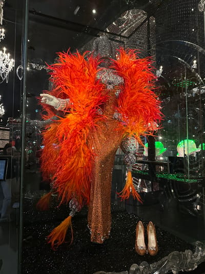 A dazzling display is presented within a glass enclosure at the Swarovski Kristallwelten in Wattens, Austria. The focal point is a mannequin adorned in an elaborate, shimmering orange gown, encrusted with countless crystals that catch the light. The gown features a dramatic, voluminous collar of bright orange and red feathers that extend outwards, framing the upper body. Two bejeweled, spherical ornaments hang from the sides of the mannequin, also sparkling with crystals. A single white-gloved hand, accented with a crystal bracelet, is extended from the mannequin's left side. At the base of the display, on a black, glitter-covered surface, rests a pair of small, sparkling orange ballet flats, each embellished with a crystal bow and the word "Paris" inscribed on the sole. The background of the enclosure is a dark, reflective surface, with hints of other crystal displays and lighting visible, including a chandelier on the left and other green crystal objects further back. Two figures are partially visible through the glass on the left side of the enclosure, one appearing to be a woman looking at an information plaque.
