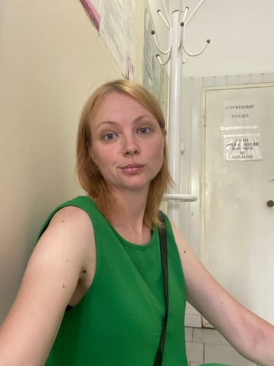 A young woman with blonde hair and blue eyes is pictured in a hallway. She is wearing a green tank top and looking at the camera. She has a small mole on her left arm. Behind her, a white coat rack stands next to a closed door with a sign that reads "СЛУЖЕБНЫЙ ТУАЛЕТ" (Service Toilet). The walls are painted a light color, and the floor is tiled. The overall setting appears to be a public building, possibly a medical clinic or administrative office, located in a city within Russia, specifically in the Zapadny district.