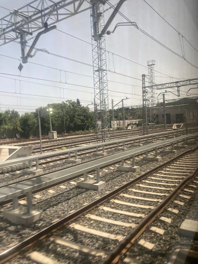 The image depicts an outdoor view of a train station with multiple sets of railway tracks in Montcada i Reixac, Spain. The scene is dominated by the infrastructure of the railway system, including overhead power lines supported by tall metal pylons, and metal safety barriers running alongside some of the tracks. The tracks themselves are made of gravel and sleepers, with steel rails. In the foreground, a section of a modern platform or walkway with a metal grating surface is visible. To the left, a white structure with the word "llaleco" printed on it is present. In the mid-ground, there is a lamppost with several lights, and beyond that, a dense line of trees forms a green backdrop. Further in the background, there is a low-rise building with windows and a hilly landscape with scattered houses. The sky is bright and hazy, suggesting daylight. There are no people visible in the image.
