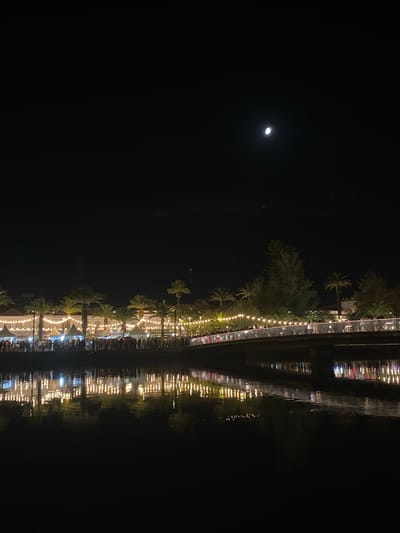 An nighttime scene captured from across a body of water shows a vibrantly lit promenade lined with palm trees and tents, likely stalls or food vendors. A bridge spans the water in the foreground, with a crowd of people visible on it and along the promenade. The lights from the promenade are reflected in the dark water. Above, the sky is dark with a crescent moon visible. The scene is situated in the city of Tavira, Portugal, likely along the Gilão River or a similar waterway.