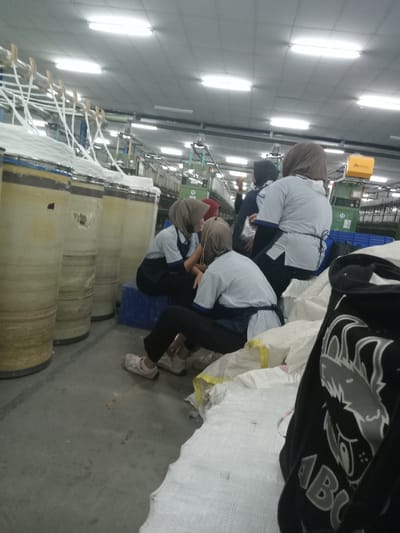 The scene is an indoor industrial facility, identified as located in Sukomoro, Indonesia, likely a textile or yarn production plant. The environment features a concrete floor and a ceiling with numerous linear fluorescent light fixtures.
Five women, appearing to be factory workers, are present. All wear head coverings (hijabs) and a uniform of light-colored shirts with dark aprons or lower garments. In the mid-ground, three women are clustered: two are seated on the floor, one wearing beige sneakers, facing each other, while a third sits on a blue plastic crate behind them. A fourth woman in the background wears a dark hijab and is seen with her right arm raised, possibly gesturing. A fifth woman with a light brown hijab is partially visible further back.
On the left, multiple large, beige, cylindrical spools or containers, banded with dark stripes and filled with white fibrous material, are arranged on the floor. To the right, a pile of white woven sacks, some with yellow markings, forms a low mound. A black garment with a grey and white graphic depicting a creature's head and the letters "ABG" is draped on these sacks in the extreme right foreground. In the background, industrial machinery with green frames and a yellow box-like component is visible.