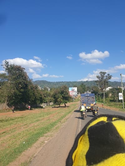 The image captures a daytime road scene in Machakos Central ward, Kenya. A clear blue sky with scattered white cumulus clouds dominates the upper portion. The perspective is from a low vantage point, looking forward over a yellow and black striped object partially visible in the bottom right corner, resembling a glove or vehicle part.
An asphalt road extends from the mid-ground towards the background. On this road, a dark blue truck with a slatted, open cargo bed is visible in the mid-distance, traveling away from the camera. Following the truck are two motorcycles. The lead motorcycle is ridden by an individual wearing a reflective vest. The second motorcycle carries a rider and a passenger, with an item secured on the rear rack.
To the left of the road, a wide, unpaved grassy verge is present, featuring multiple trees with green foliage and a low stone wall or fence line. Three human figures are discernible on this verge: one wearing a red top, one in white, and a third indistinct figure further back. To the right of the road, a similar vegetated verge includes utility poles with overhead lines. In the background, rolling green hills rise, interspersed with distant buildings. Two billboards are visible: a larger one on the left side of the road further back, and a smaller one on the right.