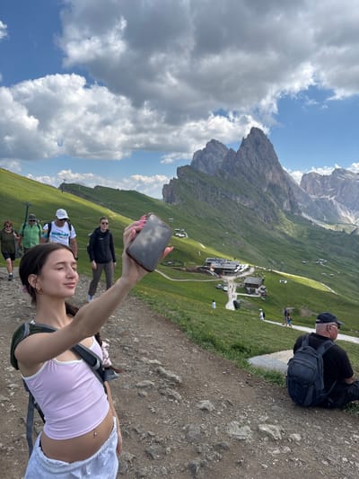 In Santa Cristina Gherdëina - St. Christina in Gröden - Santa Cristina Valgardena, Italy, a young woman is taking a selfie with her phone while on a mountain trail. She is wearing a pink tank top, white shorts, and a backpack. Behind her, other hikers are walking up the grassy slope. In the background, dramatic mountain peaks rise under a cloudy sky, and a cluster of buildings, possibly a mountain lodge or restaurant, is visible on the mountainside. The terrain is rocky with green grass and scattered yellow wildflowers.