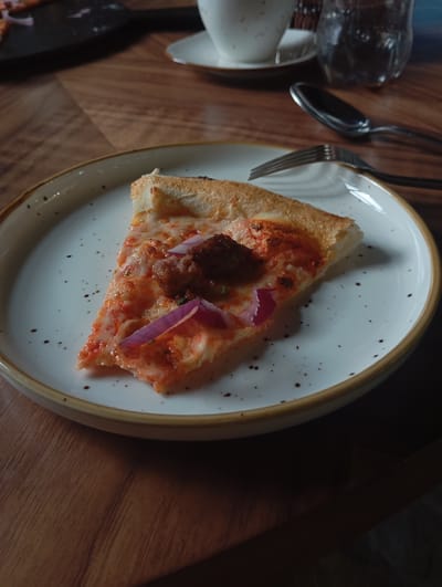 A single slice of pizza with red onion and sausage is presented on a white plate with brown speckles and a tan rim. The plate is situated on a wooden table. In the background, a white teacup with a saucer, a glass of water, and cutlery are visible, suggesting a dining setting. The lighting casts shadows, indicating an indoor environment possibly in Бактуу-Долоноту, Kyrgyzstan.