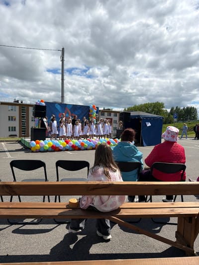 The image, taken in the city of Valozhyn, Belarus, depicts an outdoor event. A group of young people, presumably dancers, are performing on a stage decorated with balloons and a backdrop featuring a graphic image. The stage has speakers positioned in front. Several people are watching the performance, seated on chairs. The scene is partially viewed from behind a wooden structure. The weather is overcast. A building is visible in the background.