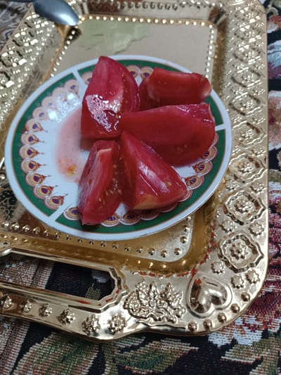 A close-up, overhead shot shows several wedges of ripe red tomatoes on a small, decorative plate. The plate has a white base with a green border featuring a repeating pattern of colorful, stylized peacock feathers. The tomatoes are glistening with juice, and some of the juice has seeped onto the plate. The plate rests on a golden, ornate tray with intricate embossed designs, including floral motifs and a heart shape. The tray itself sits on a patterned fabric that appears to be a tablecloth or runner with earthy tones and geometric designs. A silver spoon is partially visible in the upper left corner, resting on the tray. The scene is set indoors, likely in a home in the city of Balkanabat, Turkmenistan, given the style of the serving ware and the typical presentation of food in the region.