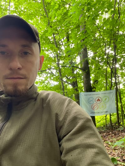 The image is a selfie taken by a person in a wooded area. The person is wearing a camouflage baseball cap and a green jacket. In the background, a white banner is hanging from a tree. The banner has several circular emblems and text in Russian. The text on the banner reads "ФЕДЕРАЦИЯ СПОРТИВНОГО ТУРИЗМА Г. САРАПУЛ" and "«ТУРИЗМ ДЕТЯМ»". The surrounding environment is characterized by lush green trees and foliage, suggesting an outdoor natural setting. The lighting indicates it is daytime, with sunlight filtering through the leaves. The specific location is identified as Nikolayevka, Russia.