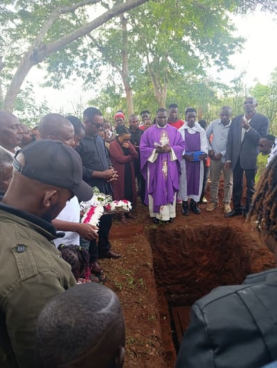 The image captures an outdoor burial ceremony in Muthetheni ward, Kenya. Approximately twenty-five individuals are gathered around a freshly dug, rectangular earth grave, which contains a visible wooden coffin at its base.
Central to the scene, a male priest, identifiable by his purple chasuble worn over a white alb, stands facing the grave. He holds a small, light-colored object, likely religious texts, in his clasped hands. To his immediate right, another male religious figure, wearing a white alb with a blue or purple stole, holds a blue container.
Attendees of various ages surround the grave and the religious figures. Many individuals wear dark, semi-formal attire. Expressions and postures vary, with several individuals appearing solemn, some with bowed heads or clasped hands. One woman to the left of the priest has her hands near her face.
In the left foreground, a man wearing an olive-green jacket and a dark baseball cap looks down towards the grave. Adjacent to him, another individual holds a floral arrangement shaped as a cross, composed of white and red flowers, positioned directly above the grave opening. Children are interspersed among the adults.
The ground consists of reddish-brown soil with sparse green vegetation. The background features numerous mature trees with green foliage, indicating an outdoor, natural environment under an overcast sky.