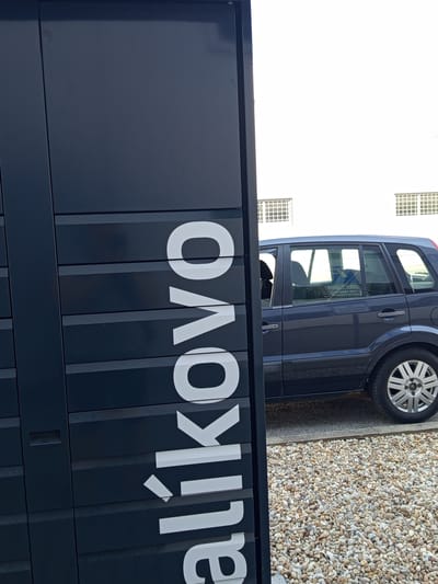 The image shows a dark blue van parked on a gravel surface next to a white building with grid-patterned windows. The van is positioned diagonally in the frame, with its rear wheel visible and partially obscured by a dark, reflective surface with large white lettering. The lettering appears to spell "OVO" followed by partially visible letters, possibly reading "alikovo" or a similar word. The context provided indicates the location is Zvolen, Slovakia. The lighting suggests it is daytime, but no specific time or weather conditions are apparent. The overall scene is static, depicting the van parked in an outdoor setting.