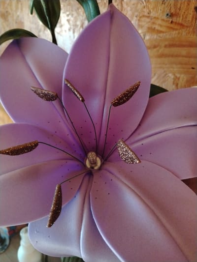 This is a close-up, medium-angle shot of a large, artificial purple lily flower. The flower is the primary subject and fills most of the frame. Its petals are soft and rounded, with subtle creases and indentations, and they have a slightly iridescent sheen. In the center of the flower, several dark green filaments extend outwards, each tipped with a textured, glittery stamen that appears to be made of a coarse, golden-brown material. Some of the stamens have a more prominent sparkle than others. The center of the flower, where the filaments originate, has a small, silver-toned, circular detail. Small, dark speckles are scattered across the surface of the petals, adding a subtle texture.

In the background, out of focus, is a light-colored, textured surface that appears to be wood paneling. To the upper left and right of the flower, dark green, stylized leaves or stems are visible, adding a contrasting color element. There is also a hint of a white, decorative object in the lower left corner of the image.

The lighting is bright, suggesting daytime, but there are no clear indicators of time of day or weather. The overall impression is one of a decorative item, likely an artificial floral arrangement. The image is cropped to focus on the intricate details of the flower. No people or text are visible in the media file. The location context provided is Zyryanskoye, Russia.