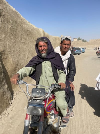 In Spin Boldak, Afghanistan, an adult male with a gray beard, wearing a dark hooded shawl over a light olive-green tunic and pants, operates a dusty, red and chrome motorcycle. His hands grip the handlebars as he looks towards the viewer. Seated behind him is a younger adult male wearing a white skullcap, a white scarf, and dark attire, with his right hand resting on the driver's back. The motorcycle is positioned on a dirt road. To the left, a tall, textured mud wall rises. The background features a wide, dusty expanse with parked vehicles, including a light blue SUV, and several individuals walking or standing. Further structures of similar mud construction are visible under a clear, bright blue sky, indicating an arid, sunlit environment.