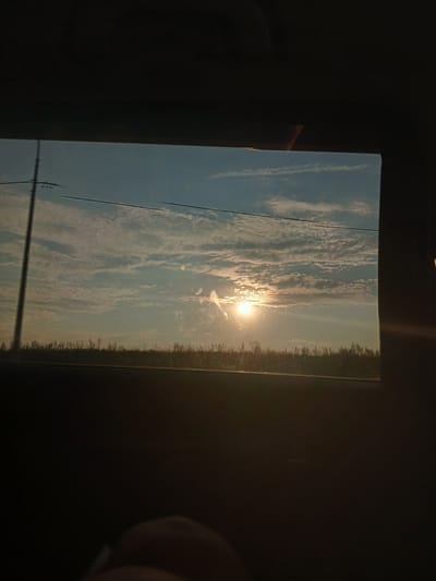 The image shows a view through a window, likely from a vehicle, looking out at a landscape. The sky is a pale blue with scattered white clouds, and the sun is visible as a bright orb, partially obscured by the clouds, casting a warm glow. A thin, dark line, possibly a telephone wire, stretches across the upper portion of the sky. In the distance, a dark, silhouetted line of vegetation or trees is visible against the sky. A vertical pole, likely a utility pole, stands on the left side of the frame, also silhouetted. The foreground and the edges of the frame are dark, suggesting the interior of a vehicle or a dark room. The bottom part of the frame shows a dark, blurry shape that appears to be part of a person's hand or arm. The overall impression is a serene, outdoor scene captured from an indoor vantage point. There are no identifiable landmarks or details to confirm the location in Teplyaki, Russia.