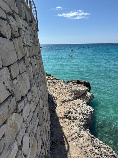 A detailed view of a coastal area in Nardò, Italy, captured on a bright, sunny day. The left foreground features a tall, light-colored stone wall composed of rough-hewn blocks, with a metal railing partially visible at its upper edge. Adjacent to the wall on the right, a narrow, uneven path of compacted earth and rock extends towards the water's edge, where a shadow is cast. The path transitions into natural rocky outcrops that meet the clear, vibrant turquoise sea. In the midground, approximately ten individuals are visible in the water. Two persons are positioned on a single stand-up paddleboard or small craft. Other individuals are distributed across the water, appearing to swim or use additional paddleboards. The background reveals the expansive deep blue sea meeting a light blue sky dotted with scattered white clouds.