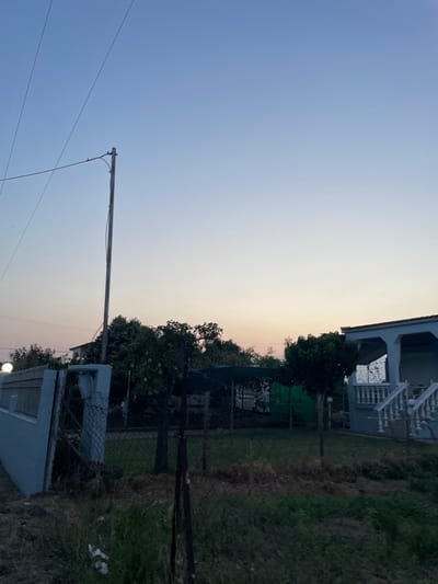 The image depicts a residential scene at dusk in Skala Avdiron, Greece. The sky is a gradient of soft blues and pale oranges, indicative of twilight. A wooden utility pole stands prominently on the left side of the frame, supporting power lines that stretch across the sky. In the foreground, a light blue wall or fence extends from the left edge of the image. Adjacent to this wall is a wire fence. Beyond the fence, a grassy yard is visible, with several trees and some covered structures. On the right side of the image, a white-columned house with a porch and a staircase is partially visible. The overall impression is a quiet, rural setting as day transitions into night.