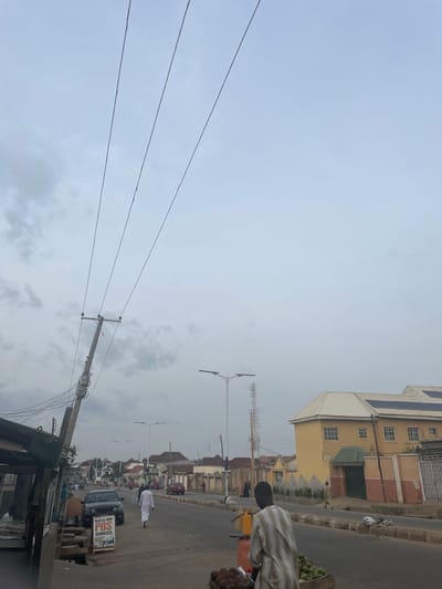 The image depicts a street scene in Unguwan Dosa, Nigeria, under an overcast sky. Power lines stretch diagonally across the frame, supported by a wooden utility pole on the left. The pole has electrical meters attached to it. A black car is parked on the right side of the road, with a sign advertising "HAJIA AL-AMIN POS SERVICES" next to it. A man in a white robe is walking away from the camera on the right side of the road, while another man, wearing a striped garment, is seen from behind in the foreground, standing near a cart laden with fruits.

Buildings line the street, with a prominent yellow structure with a white roof, featuring multiple windows and solar panels, on the right side of the road. A tall cell tower is visible in the distance. The road surface is asphalt, with some debris and puddles present. A street lamp is mounted on a pole further down the road. The environment appears to be a typical urban setting with a mix of residential and commercial structures.
