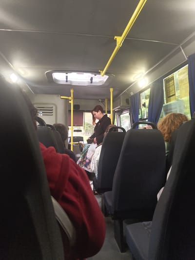 The image shows the interior of a bus in Zhukovsky, Russia, likely during daylight hours. The perspective is from a passenger seated in the back, looking forward. The ceiling of the bus is visible, with yellow handrails and lights. There are windows on the side, with blue curtains drawn. Several passengers are visible. In the foreground, parts of seats and passengers' clothing, including a red garment, are visible. Further into the bus, a woman with dark hair, wearing a black jacket, is standing near the front, looking down at something in her hands or a bag. Her posture suggests she might be retrieving or putting something away. Other passengers are seated, with their heads and shoulders visible. The environment appears to be a typical public transportation setting.