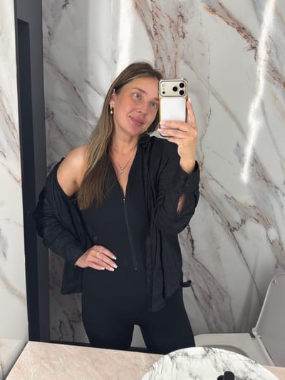 A woman is taking a mirror selfie in a bathroom. She is wearing a black jumpsuit and a black open button-up shirt draped over her shoulders. She has long blonde hair and is wearing gold hoop earrings and a gold necklace with a cross pendant. She is also wearing a gold ring with a large yellow stone on her left hand. The bathroom has marble-patterned walls and a white toilet is visible to the right. The floor in the foreground is a pinkish-white marbled countertop. The image appears to have been taken in the city of Ykremnivs'ke, Ukraine.