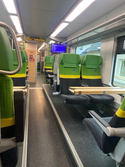 The image depicts the interior of a train carriage, likely a regional train, showing rows of seats. The seats are upholstered in a dark grey fabric with a prominent green and yellow band across the backrest. Some seats have foldable tray tables. The floor is dark grey with small white speckles. Overhead fluorescent lights illuminate the carriage. A digital display board above the seats shows "REX 6 Zielbahnhof: Bratislava-Petržalka", indicating the train's route. In the background, there is a doorway leading to another part of the train, with a sign for a "WC" (toilet). Posters are visible on a wall in the background. The overall impression is that of an empty train carriage, ready for passengers or during a quiet period. The image was taken from a low angle, looking down the aisle of the train.