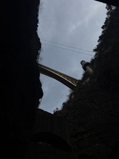 The image depicts a view looking upwards from the base of a deep, narrow gorge in Pellafol, France, under an overcast sky. Two towering, dark rock faces frame the scene, largely silhouetted, with some sparse vegetation visible along their upper edges. A modern, light-colored concrete arch bridge spans the gorge high overhead, its underside and supports visible against the sky. Below and to the left of this bridge, a section of a dark, older stone archway is visible, suggesting a lower passage or structure within the gorge. On the right side, a tall, cylindrical concrete tower structure, possibly an intake tower or part of a dam, rises from the gorge wall towards the main bridge level. Multiple thin cables or power lines traverse the gorge horizontally above the main bridge. Portions of the left rock face exhibit what appear to be protective netting or screens. No people, vehicles, or animals are visible.
