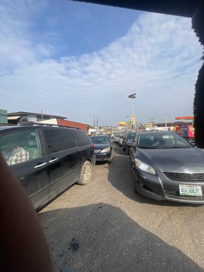 A congested street scene in Akowonjo, Nigeria, features a line of stationary vehicles under a partly cloudy sky. In the foreground, a black minivan is parked, with its side visible. To its right, a dark grey Toyota Matrix with license plate "AGL 44JY" is stopped in traffic. Beyond these vehicles, a long queue of cars stretches into the distance, navigating a paved road lined with buildings and commercial establishments, including what appears to be a gas station. The overall atmosphere suggests a typical urban traffic situation.