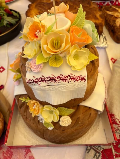 A two-tiered, braided bread loaf, likely a traditional Belarusian karavai, is presented with decorative white fondant and edible floral elements. The top tier is adorned with a bouquet of light yellow, orange, and pale green fondant flowers, some resembling roses and tulips, with delicate green leaves. A white fondant band wraps around the center of the top tier, featuring a dark red, intricate scrolled pattern reminiscent of traditional embroidery. The bottom tier is similarly decorated with smaller yellow and orange flowers and green leaves. The bread is placed in a red-lined white cardboard box, resting on a white tablecloth with a red embroidered pattern. In the background, other baked goods and a bowl of fruit are partially visible. The setting suggests a festive occasion, possibly a wedding or celebration, taking place in a home or market environment within the village of Vaynyanskiy Sel'skiy Sovet, Belarus. There are no people visible in the image.