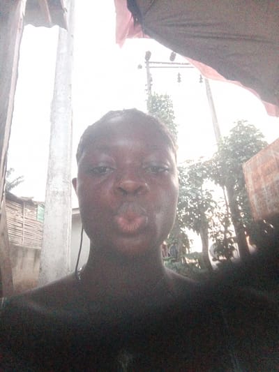 A close-up selfie appears to have been taken outside in Mbribit, Nigeria. The main subject is a dark-skinned individual with short hair, posing with pursed lips. Behind the person are trees, a wooden structure, and a utility pole, suggesting an urban or peri-urban location. The light is bright, possibly overcast, washing out some of the details in the background. A concrete post is to the person's left, and the edge of a fabric or awning appears overhead.