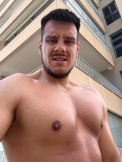 This is a first-person, low-angle, outdoor shot taken from a low perspective, looking upwards at a shirtless man. The man has dark hair and a beard, and he is smiling or grimacing with his teeth showing. He is muscular and appears to be wet, with water droplets visible on his skin, especially on his chest. His chest is the primary focus of the image, with a prominent pectoral muscle and a nipple clearly visible.

The background consists of a modern building with a beige facade and horizontal white railings or louvers. The building appears to have multiple stories, with dark-colored windows or vents visible at the top of the frame. The overall impression is that the photo was taken on a sunny day, possibly near a pool or at a resort, given the man's attire and wet appearance. The specific location context provided is Roquetas de Mar, Spain. There is no visible text in the image. The man's expression could be interpreted as one of exertion, excitement, or playful discomfort.