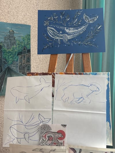 The image depicts an indoor scene in Tayshet, Russia, showcasing various artworks. Dominating the center is a wooden easel holding a horizontal blue poster featuring a stylized white whale adorned with intricate patterns and surrounded by white and black botanical elements, including leaves and star-shaped flowers. To the left, a vertical oil painting portrays a grey city or townscape under a cloudy sky, with a prominent multi-domed building resembling a church or cathedral situated on a green, tree-covered hill, and other architectural structures below. Spread across a surface in the foreground are several pieces of paper. These include multiple blue pen outline sketches of whales, a blue pen sketch of a quadruped animal resembling a bear, and a detailed monochromatic illustration of a patterned creature, possibly a badger, against a red circular background. The background behind the easel and paintings consists of a textured, light-colored wall and vibrant turquoise curtains on the right. A light-colored, possibly carpeted, surface is visible at the bottom of the frame. No individuals are present.
