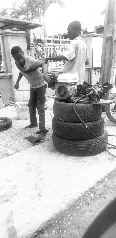 In Kinkinau, Nigeria, two young boys are engaged in mechanical work outdoors. The scene appears to be a makeshift workshop. The boy on the left, wearing a t-shirt and jeans, is bending over and appears to be adjusting a piece of equipment. His bare feet are visible, and he is barefooted. The boy on the right, in a button-up shirt and jeans, is standing and also interacting with the equipment. The primary object of their attention is a small engine, possibly for a generator or pump, which is mounted on a stack of three tires. A black hose is connected to the engine and extends downwards. In the background, a metal fence and buildings are visible, along with palm trees, suggesting a tropical or sub-tropical urban environment. The ground is paved with concrete, and there are various discarded items scattered around, including a bucket, a tire, and a piece of black material. The overall atmosphere suggests a practical, hands-on approach to repair or fabrication in an open-air setting.