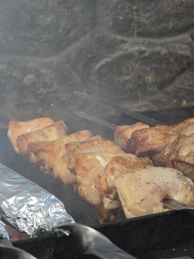 The image shows a close-up view of meat skewers cooking on a grill, possibly in Pidhorodne, Ukraine. The meat, appearing to be chicken or pork, is browned and seasoned with pepper. It is threaded onto metal skewers and arranged horizontally on the grill. Smoke is visible, indicating the cooking process. In the foreground, a skewer is partially wrapped in aluminum foil, and a metal utensil is also visible. The background consists of a rough, textured stone surface, which could be part of a grill enclosure or a decorative element. The scene is dimly lit, with the focus on the food.