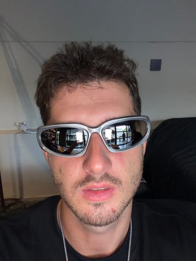 A close-up, head-and-shoulders portrait features a male individual with short, dark brown hair and light facial stubble. The subject wears silver-framed, reflective, wraparound sunglasses, a black t-shirt, and a silver-colored chain. A minor blemish is visible on the subject's chin and lower lip.
The reflections within the sunglasses reveal an indoor environment located in Tumbak Bayuh, Indonesia. The reflections show a spacious area characterized by large, dark-framed windows or glass doors. In the left lens, a person's arm and hand are visible, potentially holding an object, alongside a wooden surface and a blurred individual. The right lens reflects a bright rectangular object, an additional standing figure, and further window elements.
The background directly behind the subject consists of a plain, light-colored wall. Subtle vertical shadows, consistent with blinds, are visible on the upper left wall section. A small, dark blue square object is affixed to the upper right wall. A white fixture is partially visible on the left side, near the subject's left shoulder. An out-of-focus dark object occupies the right edge of the frame.
