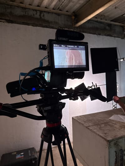 The image depicts an indoor film or video production setup in El Paraíso, Mexico. A professional video camera, equipped with a RØDE shotgun microphone and an external monitor, is centrally positioned on a black tripod with red accents. The monitor displays a close-up of a subject with pink or reddish hair. Various cables, including a coiled blue one, connect components on the camera rig. To the right of the camera, a dark, rectangular object is mounted on a stand or attached to the wall, possibly a light panel or flag, with a bright light source illuminating the background behind it. A light-colored wooden cabinet or table is located in the lower right foreground. Partially visible on the far right is a human hand holding a light-colored object, possibly a prop or tool. The room features light-colored walls and an unfinished ceiling with visible joists and possibly corrugated material. A black hard-shell case, labeled "PELICAN," rests on the floor in the lower-left corner.