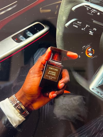 A person's hand, with dark skin and orange-painted fingernails, holds a bottle of Tom Ford Myrrhe Mystère Eau de Parfum. The bottle is dark brown glass with a silver cap. The person is wearing a silver and rose gold watch on their wrist. The background appears to be the interior of a car, with a steering wheel visible on the left side of the image. Buttons for cruise control are labeled "OK", "RES/SET+", "CANCEL", and "SET-". A circular gauge with orange and black markings is also visible. The overall lighting suggests it is daytime, but the exact time cannot be determined. The setting is likely a car interior in Borowa, Poland.