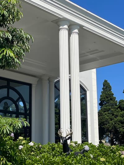 The image presents an exterior view of a building in Kayuambon, Indonesia, characterized by classical architectural elements under a clear blue sky. The structure is predominantly white, featuring several large fluted columns. Two prominent columns occupy the central upper mid-ground, supporting a broad, flat white overhang with visible recessed lighting fixtures. To the left, a section of the building's facade includes large, dark-framed windows with distinctive arched tops, reflecting surrounding greenery and the sky. Further columns are partially visible to the left and right sides of the building.

The lower portion of the scene is dominated by dense, vibrant green foliage, interspersed with clusters of light purple and white flowers. Within this greenery, a black statue of a deer with light-colored antlers is prominently positioned in the lower-center. A portion of a second black deer statue is visible to its right, also nestled within the bushes. A tree with broad green leaves extends into the upper left quadrant, partially obscuring the building's facade. In the background, on the right, several tall, dark green coniferous trees stand against the bright blue sky. The scene is illuminated by strong daylight, casting defined shadows. No people are present.