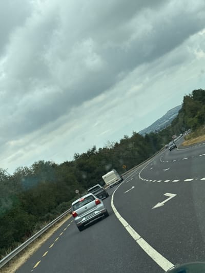 The image shows a view from inside a vehicle on a highway, looking ahead and slightly to the right. The sky is overcast with thick, gray clouds. The road curves to the right, with multiple lanes marked by white lines and directional arrows indicating a left turn. Several vehicles are visible on the road, including a silver Volkswagen Polo, a white caravan being towed, and other cars further in the distance. The road is bordered by a metal guardrail on the left and a densely wooded hillside on the right. Distant hills and buildings can be seen in the background. The camera angle is tilted, suggesting the image was taken while the vehicle was in motion. The location is identified as Kilivory, Ireland.