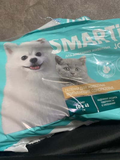 A close-up, angled shot shows a package of pet training pads. The packaging is teal and white with illustrations of a white dog and a grey cat. Text on the package is in Russian and includes brand names and product details. The package appears to be placed on a dark, textured surface, possibly a bag or a piece of furniture. The background is out of focus and shows a rough, textured wall. There is no visible indication of the location being in Akhtyrskiy, Russia, or any other specific city.