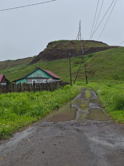 An exterior, eye-level, medium shot captures a rural scene in Uglegorsk, Russia. The focal point is a narrow, muddy dirt road leading away from the viewer, lined with lush green grass and dotted with large puddles reflecting the overcast sky. To the left, a small, weathered wooden house with a red roof and green siding stands behind a rustic wooden fence. Beyond the house, another structure with a red roof is partially visible. In the background, a steep, grassy hill rises, with visible erosion and dark soil patches. Power lines crisscross the sky, supported by wooden poles. The overall atmosphere is one of dampness and quiet solitude.