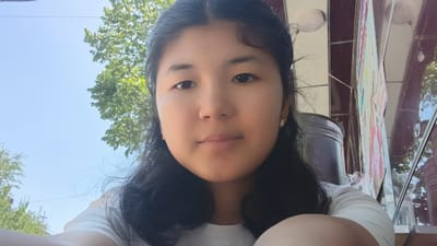 A young woman with dark hair pulled back is looking directly at the camera in a selfie. She has brown eyes and is wearing a white t-shirt and small gold earrings. Her face is softly lit, suggesting it is daytime. Behind her, a bright blue sky is visible with the leaves of a tree to her left, indicating an outdoor setting. To her right, part of a building with a dark red facade and some colorful signs or posters is visible. The overall scene appears to be in Osh City, Kyrgyzstan, on a clear day.