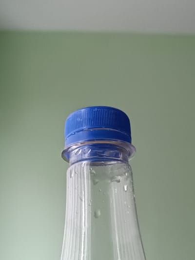 The image shows a close-up, low-angle shot of the top of a clear plastic bottle with a blue screw-on cap. The bottle has vertical ridges and some water droplets clinging to its surface. The background is a solid, light green wall. There is no indication of the location being in the city of Ключи, Russia, or any other specific geographical place. The scene is static and focuses solely on the bottle and its immediate surroundings.