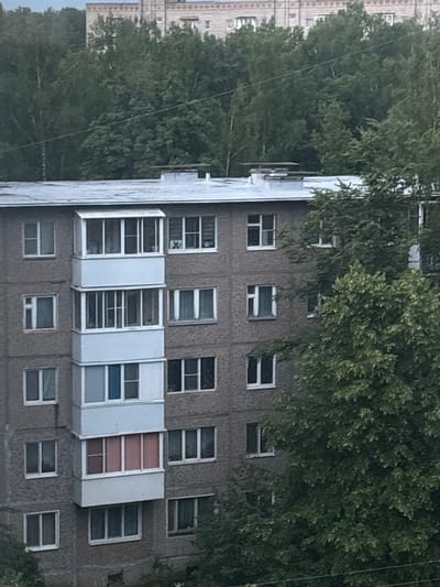 The image shows a grey, multi-story apartment building with a balcony on the front facade. The building has numerous windows, some of which are open. Trees are visible in front of and behind the building, and a larger building with white trim is visible in the background. The sky is overcast. The location is identified as Radkevshchina, Russia. There are no people visible in the image.