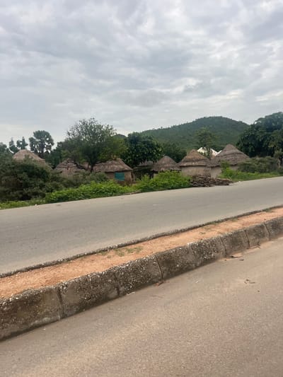 A paved road runs diagonally across the foreground of the image. The road is bordered by a concrete curb with a reddish-brown gravelly surface. In the background, a cluster of thatched-roof huts with mud walls are visible, surrounded by lush green foliage and trees. Behind the huts, a verdant, rolling hill rises under a cloudy, overcast sky. The scene appears to depict a rural or semi-rural environment in Kona Garu, Nigeria.